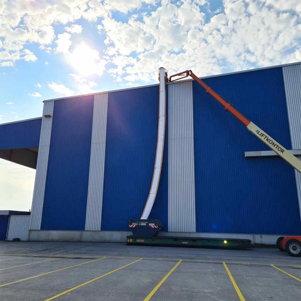 PP rubble chute raised overhead by a boom lift inside a blue-painted factory hall