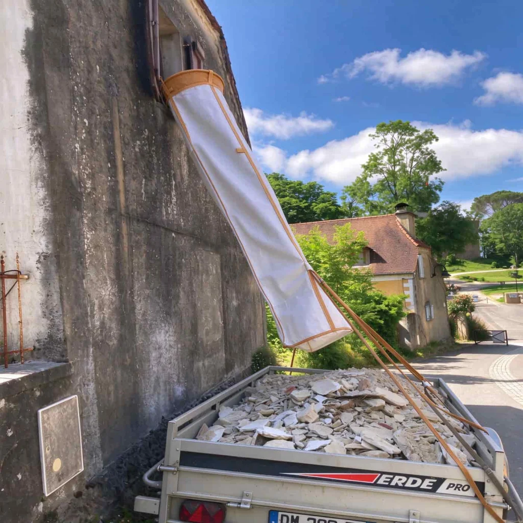 PP rubble chute hanging at a stone-wall site, slung from the trailer on its camel webbing strap